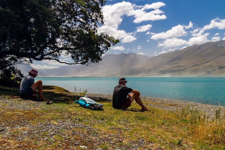 2 people sitting at the shore of Lake Ohau watching dust blowing up in the distance