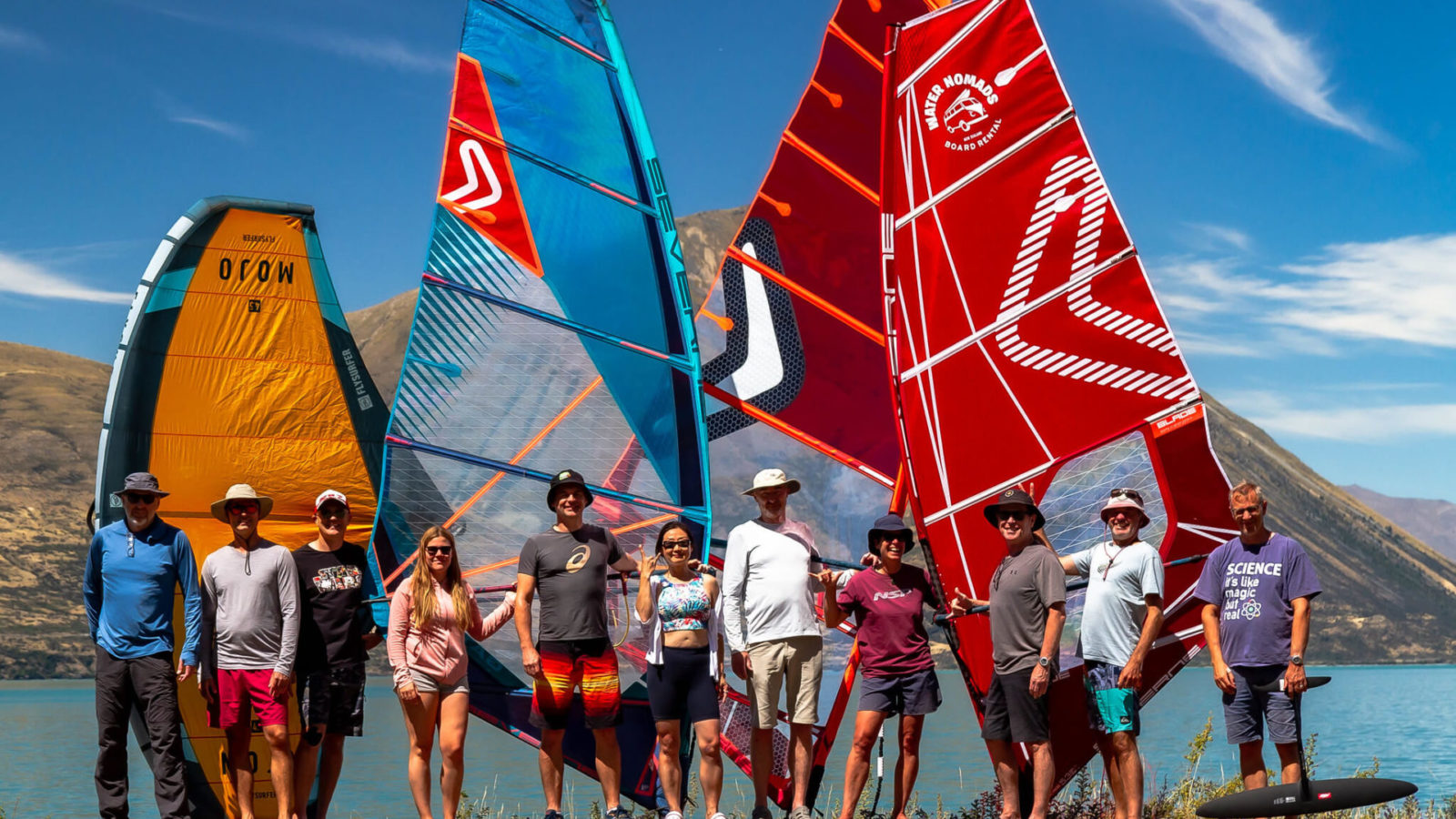 11 windsurfers posing in front of some sails with lake and mountain backdrop